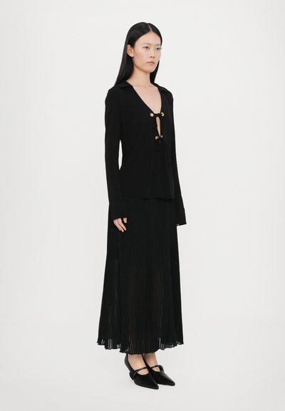 Black long-sleeve top with a deep V-neck and gold buttons, paired with a black pleated skirt that flows to the floor and pointy black shoes.