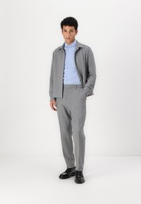 Traje gris a rayas con una camisa abotonada en azul claro. Incluye un abrigo con cuello y pantalones a medida, combinados con zapatos negros.