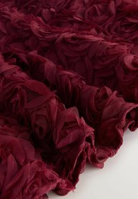 Burgundinės gėlių medžiagos, kurioje pavaizduoti sluoksniuoti, persidengiantys žiedlapiai iš lengvo tinklelio, sukuriantys tekstūruotą, trimatį vaizdą.