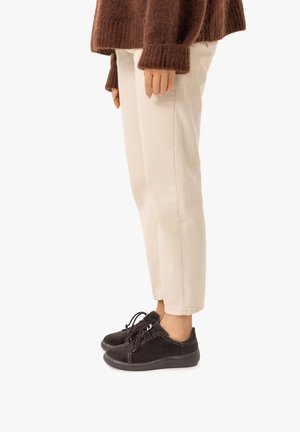 Persona che indossa pantaloni beige, un maglione marrone e scarpe sportive nere con lacci, in piedi davanti a uno sfondo bianco semplice.