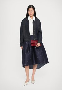 Woman wearing a dark denim jacket, white collared shirt, navy floral midi skirt, white heels, holding a red clutch bag.