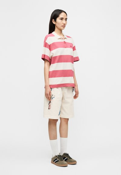 Polo rayé rouge et crème, mélange de coton, manches courtes. Shorts beige avec broderies colorées. Chaussettes blanches jusqu'aux chevilles et baskets vertes.