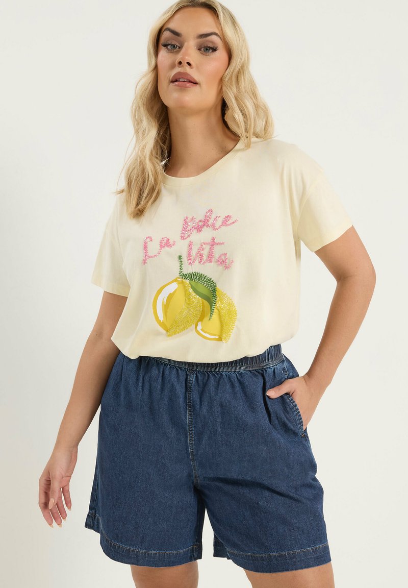 Femme aux longs cheveux blonds portant un t-shirt crème avec des citrons et le texte « La dolce vita », associé à un short en denim bleu foncé.