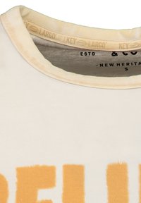 Camiseta crema con un escote redondeado. Presenta grandes letras en bloque naranjas. Hecha de una tela suave y elástica. La etiqueta muestra la marca y la talla.