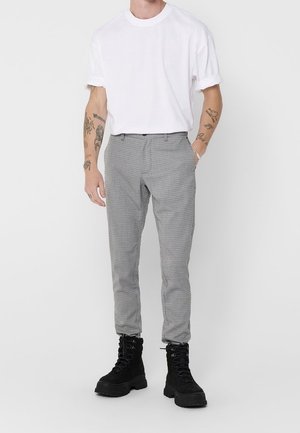 Homme portant un t-shirt blanc uni à manches retroussées, un pantalon slim à carreaux gris et des bottes de combat noires à lacets, debout avec une main dans la poche.
