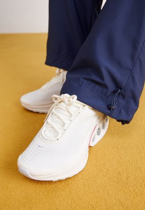 Białe sportowe trampki z siateczkowymi cholewkami noszone z granatowymi spodniami na dywanie w kolorze musztardowej żółci.