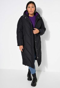 Svart puffjacka med huva, med quiltad struktur och silverfärgad dragkedja. Bärs över en lila tröja, i kombination med blå jeans och svarta stövlar.