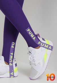 Białe buty sportowe z purpurowymi akcentami i żółtym detalem na podeszwie, zestawione z dopasowanymi, prążkowanymi purpurowymi legginsami z logo "PUMA".