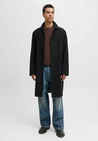 Zwarte overjas met een recht, lang ontwerp, gedragen over een bruine trui en losse blauwe jeans. Voorzien van grote knopen en zijzakken.
