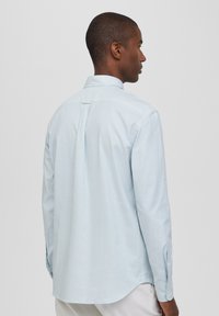 Camicia a righe blu chiaro con colletto standard e maniche lunghe. Presenta una texture sottile e un orlo arrotondato. Bottoni sui polsini.