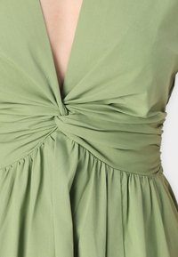 Abito verde con profondo scollo a V, dettaglio frontale intrecciato e gonna arricciata. Realizzato in tessuto liscio con una leggera texture.