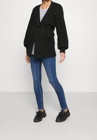 Svart stickad cardigan med bälte, ribbad textur, vida ärmar, använd över en randig topp och kombinerad med smala blåjeans och svarta skor.