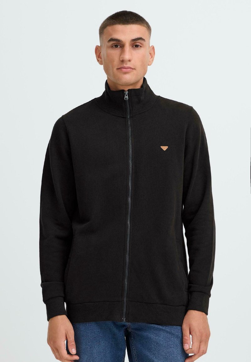 Sudadera negra con cremallera, cuello alto, puños y dobladillo de canalé. Presenta un pequeño parche de logo marrón en el pecho. Textura de tela suave.