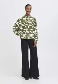 En kvinde står smilende iført en grøn og hvid mønstret bluse med knytning i halsen og sorte brede bukser foran en ensfarvet hvid baggrund.