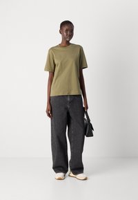 Olivgrünes Baumwoll-T-Shirt, kurze Ärmel, lockere Passform. Kombiniert mit großen schwarzen Jeanshosen und Sneakers mit Creme- und Beigetönen.