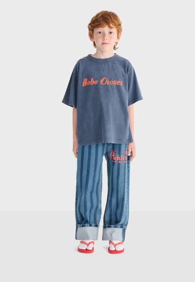 Kind met rood haar dat een losse marineblauwe "Bobo Choses" T-shirt draagt, een brede gestreepte spijkerbroek met rode tekst, en rode teenslippers met witte sokken.
