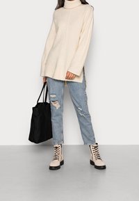 Beige oversized turtleneck sweater, distressed light blue jeans, and cream lace-up boots with black soles. Black tote bag in hand.