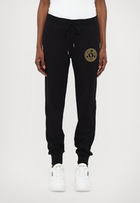 Svart bomullsjoggebukser med elastisk midje, snøring og en gullfarget sirkellogo på venstre hofte. Fitted mansjetter ved ankelen. Hvite sneakers.