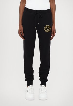 Svart bomullsjoggebukser med elastisk midje, snøring og en gullfarget sirkellogo på venstre hofte. Fitted mansjetter ved ankelen. Hvite sneakers.