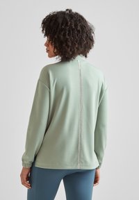 Green sweat top with a high collar, back zipper detail, and long sleeves, made of smooth fabric. Slightly loose fit, hem ends at hip.