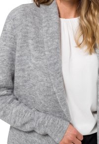 Cardigan grigio in maglia con una texture morbida, davanti aperto e maniche lunghe. Indossato sopra una camicia bianca, con un motivo a coste sui polsini.