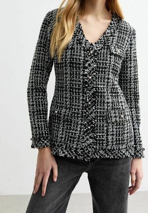 Mujer con chaqueta de tweed blanca y negra con botones de perlas, de pie con una mano en la cintura, combinada con pantalones gris oscuro.