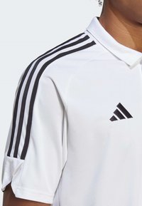 Polo bianco con tessuto a trama, con strisce nere sulle spalle e il logo Adidas in nero sul lato superiore sinistro.
