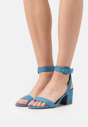 Sandales block-heel en suède bleu avec une large lanière sur les orteils et une lanière ajustable à la cheville. Texture lisse, talon bas, design minimaliste.