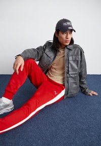 Grå skinnjakke med flere lommer, tan t-skjorte, røde joggebukser med hvite striper, grå sneakers og en mørk caps.