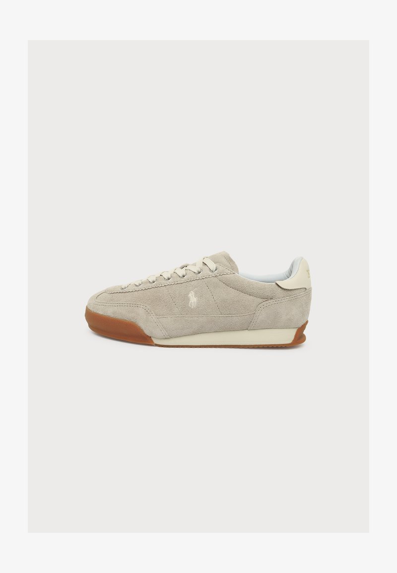Baskets en daim beige avec des lacets blancs, une semelle en gomme et un petit logo brodé sur le côté, vue de la gauche sur fond blanc.