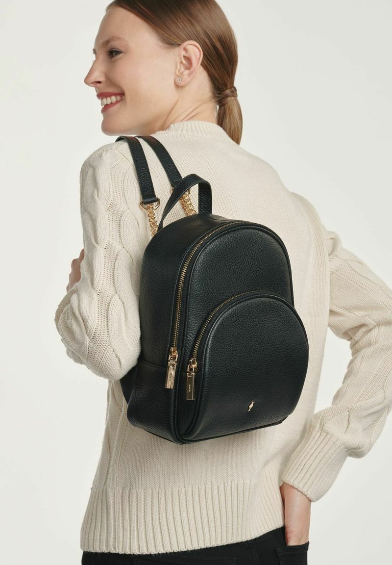 Czarna skórzana plecak o teksturowanym wykończeniu, zaokrąglonym kształcie, podwójnych zamkach i złotych akcentach. Noszony na kremowym swetrze.