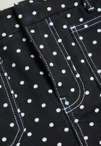 Sort stof med hvide polkaprikker, der viser nærbillede af syede samlinger og lommekanter med kontrasterende hvid tråd.