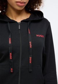 Svart hoodie med dragkedja fram, röd logotyp och snören. Har en känguruficka och en mjuk textur, designad för komfort.