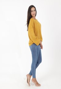 Musztardowy gruby sweter dziany z bufiastymi rękawami, zestawiony z obcisłymi niebieskimi dżinsami i przezroczystymi sandałami na obcasie, na białym tle.