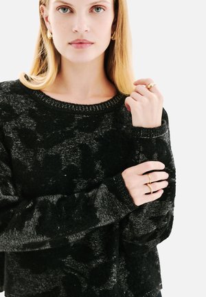 Pull en texture noire avec une coupe décontractée, doté d'un col rond et de motifs sombres contrastants. Les accessoires comprennent des bagues et des boucles d'oreilles en or.