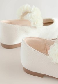 Bílé textilní boty s texturovaným povrchem, zdobené jemnými bílými květinovými detaily a nízkou hnědou podrážkou.