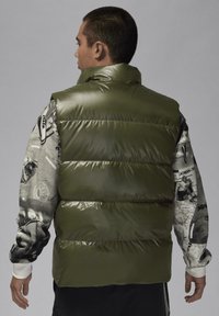 Grön pufferväst med en glansig yta, med horisontell quiltning. Modellen bär en grafisk långärmad skjorta under.