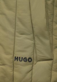 Casaco acolchoado verde-oliva com costura vertical, apresentando um logótipo preto "HUGO" no canto inferior direito e textura suave.