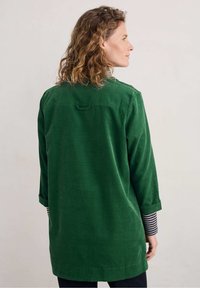 Giacca camicia in velluto a coste verde con vestibilità rilassata, caratterizzata da maniche arrotolate e un piccolo anello sulla parte posteriore del collo per appenderla. Polsini interni a righe visibili.