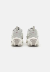 Nike lenkkarit, joissa on vaaleanharmaa päällinen, teksturoitu sivu, vaaleanpunainen logo ja paksu valkoinen pohja, jossa on voimakkaat urapatterit.