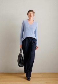 Φωτεινό μπλε ριγμένο πλεκτό πουλόβερ, σχέδιο σε V-λαιμό. Συνδυασμένο με μαύρα παντελόνια με ρίγες. Το μοντέλο κρατά μια μαύρη τσάντα tote και φοράει μαύρα παπούτσια.