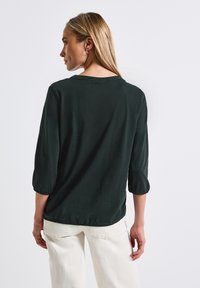 Blusa leggera di un verde scuro, con maniche a tre quarti; texture liscia e vestibilità rilassata, dettagliata con un sottile scollo rotondo.