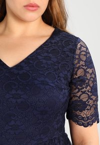Granatowa koronkowa sukienka z dekoltem w serek i krótkimi rękawami. Rękawy mają wzór kwiatowy i falbany, co nadaje fakturowany wygląd.