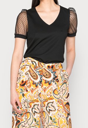 Vrouw die een zwart V-hals topje draagt met doorschijnende gestippelde pofmouwen en een hooggetailleerde wijde broek met een oranje paisley patroon.