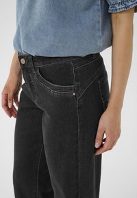 Vaqueros de mezclilla negra con corte de altura media, costuras en contraste y bolsillos frontales. Una camisa de mezclilla azul está metida en la cintura.