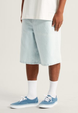 Pantalones cortos a rayas azul claro, fabricados en un tejido suave, hasta la rodilla, con un corte recto. Combinados con zapatillas de lona azules y calcetines blancos de caña baja.