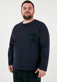 Tumesinise pikkade varrukatega särk, mis on valmistatud puuvillast ja millel on rohelise ja musta ruuduline tasku. Mudel kannab sarnaste värvidega ruudulisi pükse.