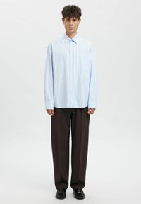 Lys blåstripet langermet skjorte med knappestolpe og én brystlomme, kombinert med løse brune bukser og svarte sko.