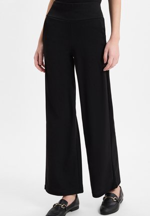 Femme portant un pantalon noir taille haute à jambes larges, associé à des mocassins en cuir noir avec des détails de boucle dorée.