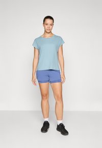 Gaiši zila tshirta ar īsām piedurknēm un purpura šortiem. Apģērbs ir brīvas formas un to papildina melni sporta apavi.